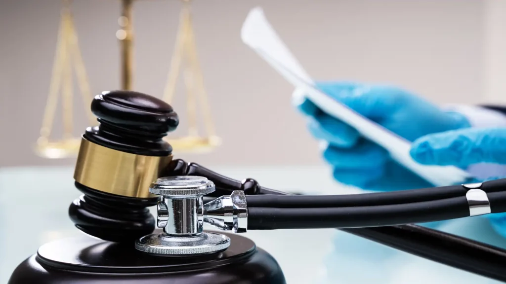 A close-up image of a gavel and stethoscope with a hand holding legal documents in the background