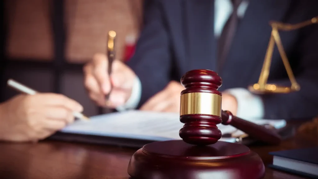 A close-up of a gavel on a table with a person writing on a document in the background, symbolizing a legal setting.