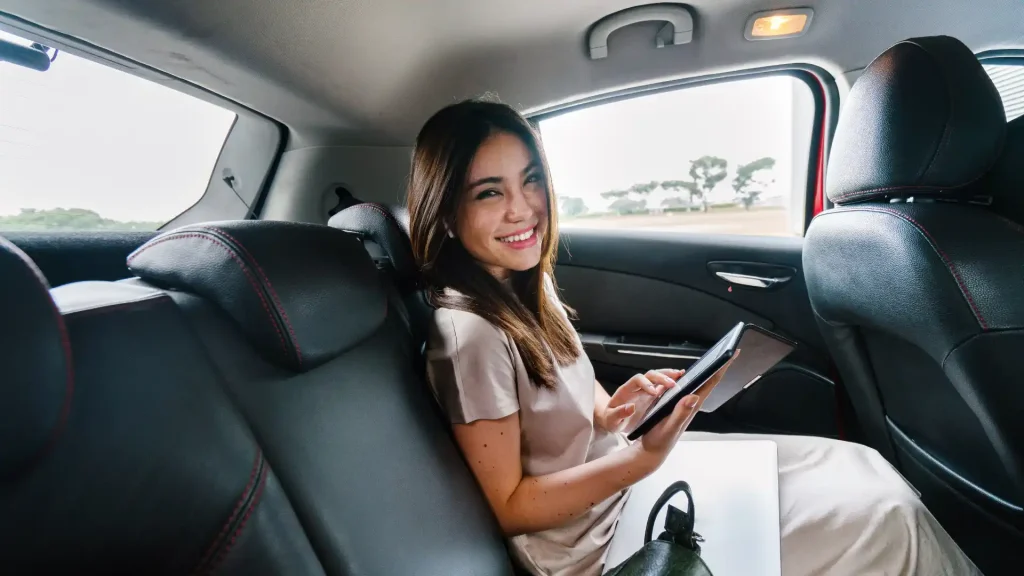 A woman sitting in the back seat of a car, smiling and using a tablet.