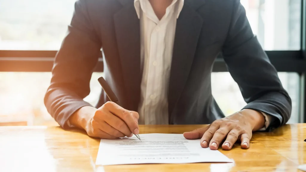 Person signing a document on a wooden table, with a pen in hand