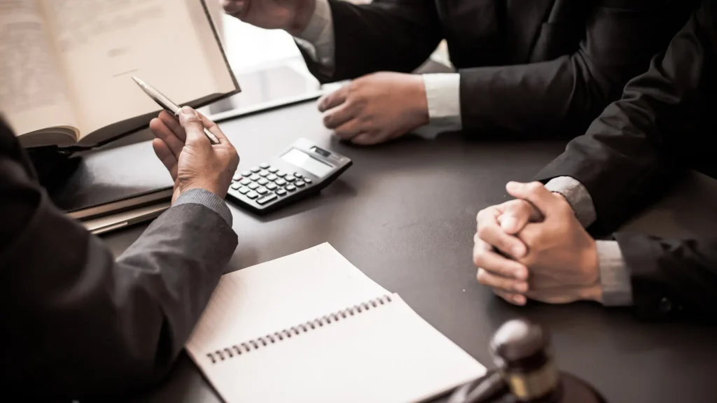 Two people in business suits discussing documents on a desk with a calculator and notepad.
